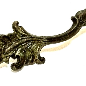 May include: Close-up of an antique-style metal drawer pull. The pull has a curved handle and ornate, leaf-like detailing. The metal has a tarnished, aged appearance, suggesting it is vintage or vintage-inspired.