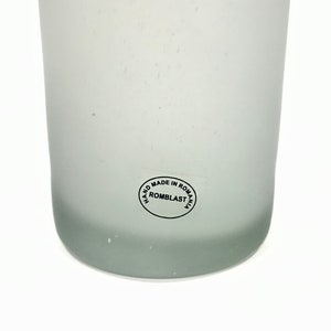 May include: A frosted glass vase with a slightly rounded base. The vase has a smooth, matte finish and a subtle gradient from clear to a light gray at the bottom. A circular sticker on the side reads "HAND MADE IN ROMANIA ROMBLAST".