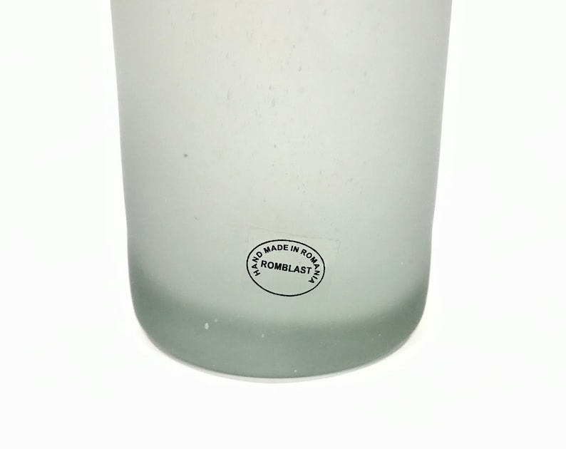 May include: A frosted glass vase with a slightly rounded base. The vase has a smooth, matte finish and a subtle gradient from clear to a light gray at the bottom. A circular sticker on the side reads "HAND MADE IN ROMANIA ROMBLAST".