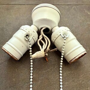 May include: A vintage white ceiling light fixture with three bulb sockets. Each socket has a pull chain with a decorative end. The fixture is mounted on a brown surface.