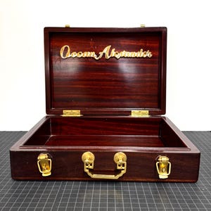 May include: A dark brown wooden briefcase with a polished finish, featuring gold-colored metal latches and a central handle. The interior lid displays the gold-colored text Ocean Alexander in a cursive script, with gold hinges connecting the lid.