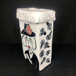 May include: A white ceramic vase with a black and orange illustration of a woman wearing a hat. The vase has a white, textured rim and is decorated with black roses. The vase is on a black surface.