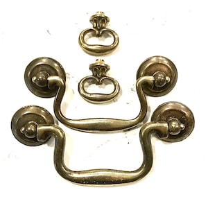 May include: Antique brass drawer pulls and knobs. The drawer pulls have a curved handle design with round backplates. Two smaller knobs and two larger pulls are arranged on a white surface. The hardware has a vintage aesthetic.