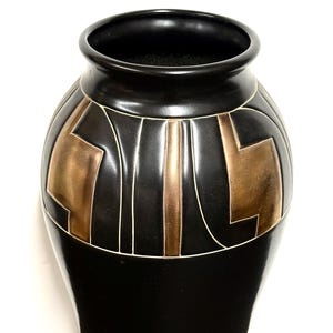 May include: A matte black ceramic vase featuring a wide, rounded body and a narrow neck. The upper portion of the vase is decorated with a series of geometric, metallic bronze-colored panels separated by thin, white incised lines.