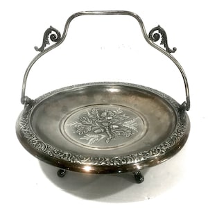May include: Silver-toned serving basket with a handle and ornate detailing. The round dish features an embossed oak tree design in the center and a decorative border. The handle has decorative scrollwork on each side. The basket rests on three small feet.