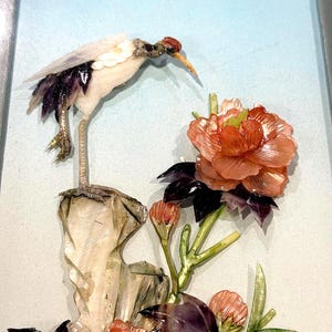 May include: A framed wall art piece featuring a crane standing on a rock, crafted from iridescent seashells. The crane has white and dark purple plumage, while a large, layered orange flower with dark purple leaves sits below it on a light blue background.