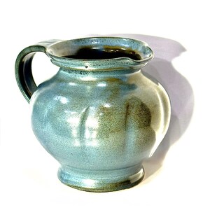 May include: A ceramic pitcher with a rounded body and a wide, flared rim. The exterior features a mottled blue and green glaze with vertical indentations along the sides. It has a single curved handle on the left side and a dark interior.