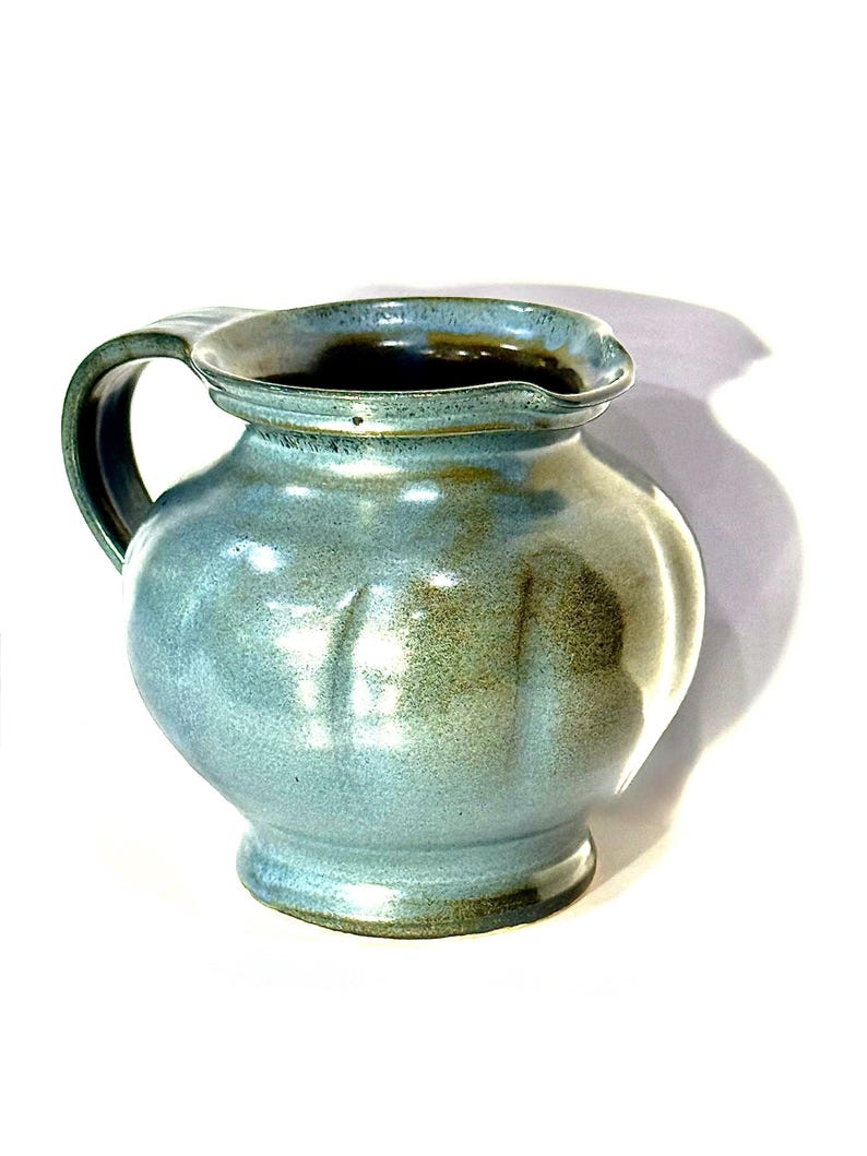 May include: A ceramic pitcher with a rounded body and a wide, flared rim. The exterior features a mottled blue and green glaze with vertical indentations along the sides. It has a single curved handle on the left side and a dark interior.