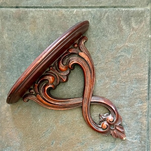 Puede incluir: Estante de pared de madera marr&oacute;n oscuro y ornamentado con una parte superior curvada de forma ovalada. El estante presenta intrincados adornos tallados y un elemento floral decorativo en la parte inferior. El estante est&aacute; dise&ntilde;ado para montarse en una pared.