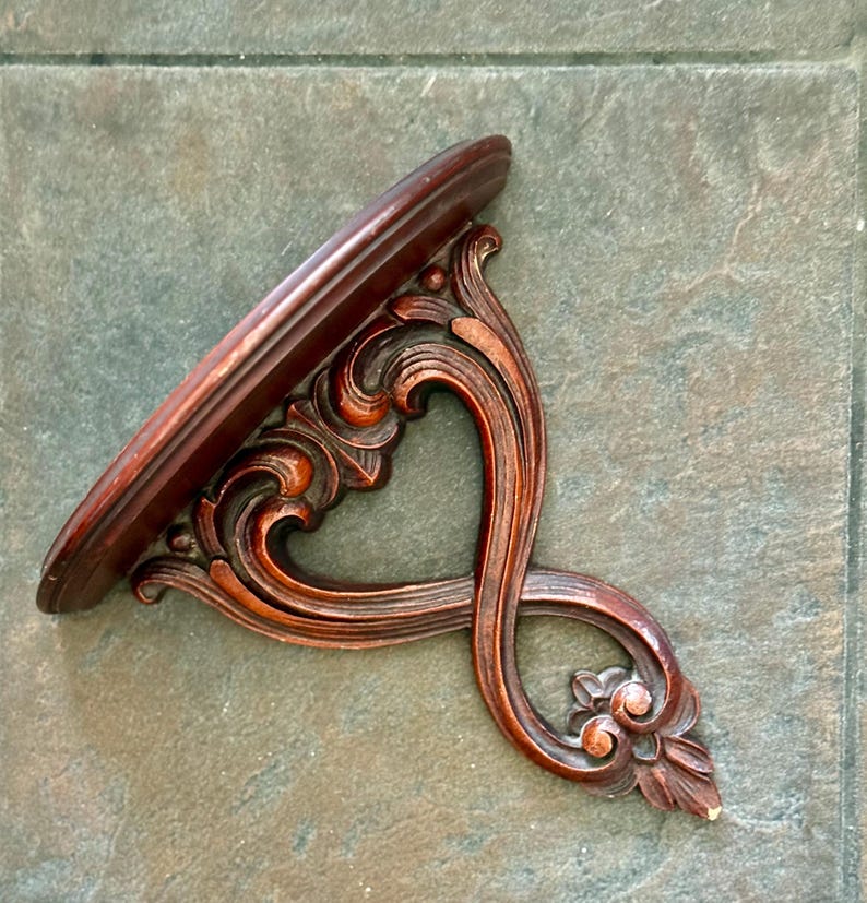 Puede incluir: Estante de pared de madera marr&oacute;n oscuro y ornamentado con una parte superior curvada de forma ovalada. El estante presenta intrincados adornos tallados y un elemento floral decorativo en la parte inferior. El estante est&aacute; dise&ntilde;ado para montarse en una pared.