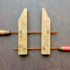 May include: A vintage parallel clamp featuring two light-colored wooden jaws and two threaded metal rods. The clamp includes one wooden handle with a worn finish and one handle painted red, both attached to the rusted metal screws for adjustment.
