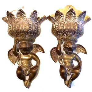 May include: Two antique brass wall sconces, each featuring a cherub holding a decorative floral element. The sconces have a warm, aged patina, with intricate detailing on the cherubs and the floral designs. The sconces are designed to be mounted on a wall.