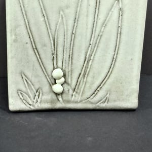 May include: A rectangular, off-white ceramic tile with a raised relief design of stylized grass and three small, round, raised accents. The tile has a slightly textured surface and a matte finish. The design is in a neutral color.