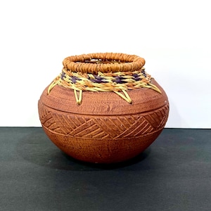 May include: A handcrafted, terracotta-colored ceramic pot with a woven rim and decorative carved geometric patterns. The rim features a woven design with natural, purple, and tan fibers. The pot is round with a textured surface.