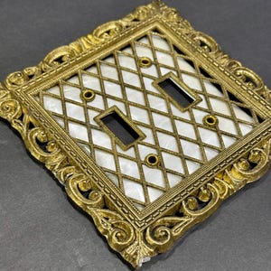 May include: Ornate, antique-style light switch cover with a gold-colored metal frame. The cover features a diamond-patterned grid over a white background, with two rectangular switch openings and decorative accents.