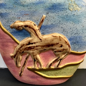 May include: A ceramic vase with a horse relief. The vase features a light brown horse against a blue and pink background. The horse is in a running position, with a gold trim around the edges. The vase has a unique, artistic design.