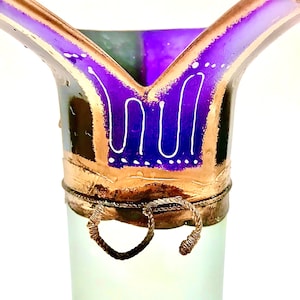 May include: A decorative glass vase with a flared top. The vase features a purple and gold rim with white accents. The body of the vase is a pale green color. Two wire handles are attached to the base of the rim.
