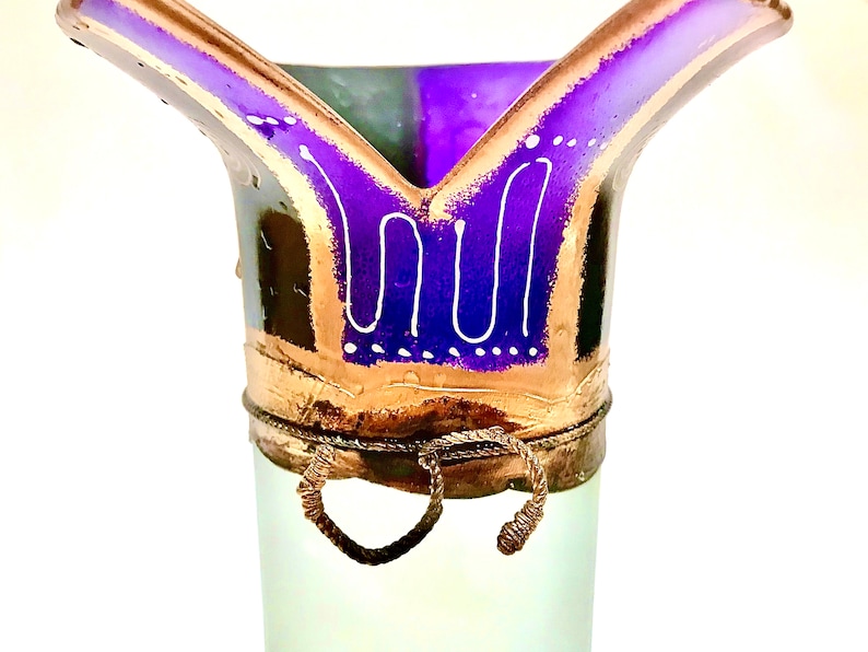 May include: A decorative glass vase with a flared top. The vase features a purple and gold rim with white accents. The body of the vase is a pale green color. Two wire handles are attached to the base of the rim.
