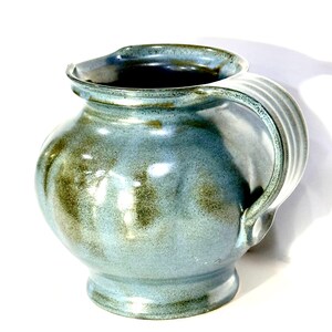 May include: A ceramic pitcher with a rounded body and a wide, flared rim. The surface features a mottled glaze in shades of light blue and earthy olive green. A sturdy, ribbed handle is attached to the side, and the interior is dark.