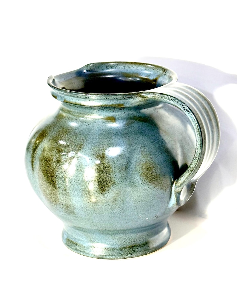 May include: A ceramic pitcher with a rounded body and a wide, flared rim. The surface features a mottled glaze in shades of light blue and earthy olive green. A sturdy, ribbed handle is attached to the side, and the interior is dark.