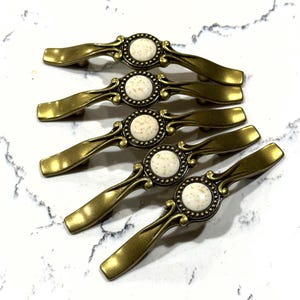 May include: Five antique brass cabinet pulls with a cream-colored center and decorative detailing. The pulls have a curved handle design and are arranged on a white marble surface. These decorative pulls are ideal for adding a vintage touch to furniture.