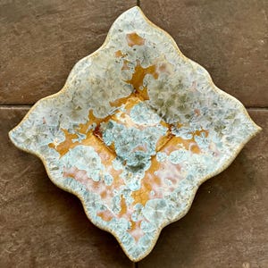 May include: A decorative ceramic dish with a unique, four-lobed shape. The dish features a blend of colors, including gold, light blue, and pink, with a crystalline pattern. The edges are slightly raised and have a textured appearance.