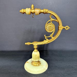 May include: A gold-toned metal display stand featuring a curved arm with ornate scrollwork and a circular floral medallion. The base and top accents are crafted from smooth, pale green onyx stone, with a small metal hook hanging from the arm.