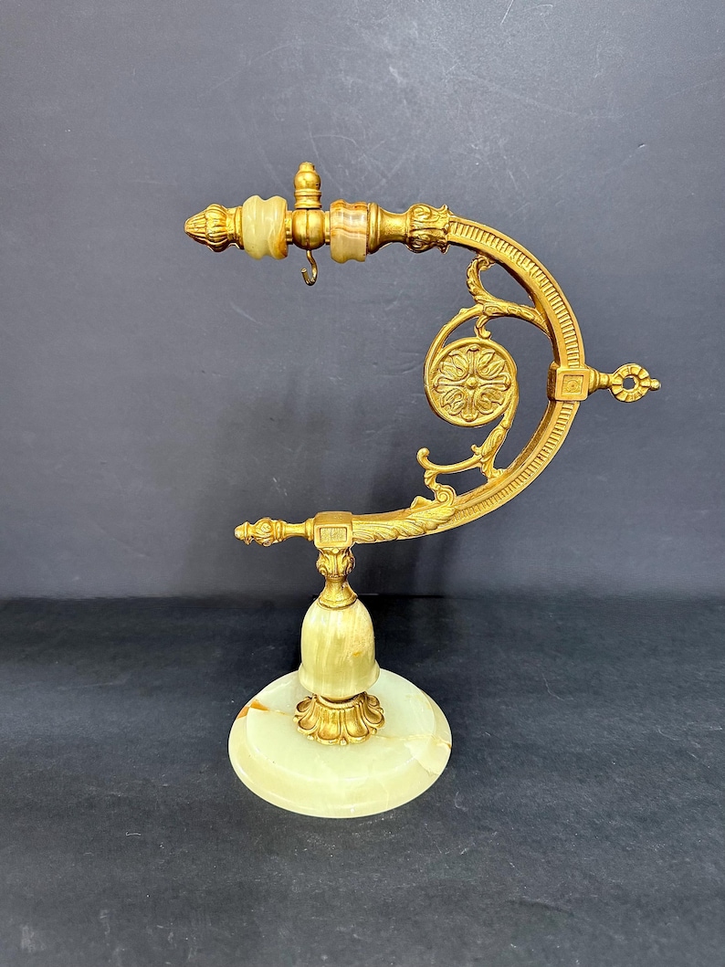May include: A gold-toned metal display stand featuring a curved arm with ornate scrollwork and a circular floral medallion. The base and top accents are crafted from smooth, pale green onyx stone, with a small metal hook hanging from the arm.