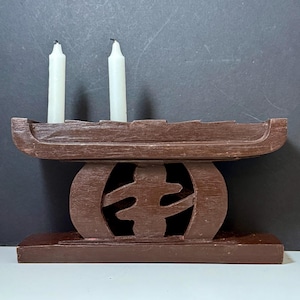 May include: A brown wooden candle holder with two white candles. The holder has a rectangular base, a decorative central element, and a top section designed to hold the candles. The design is simple and elegant.