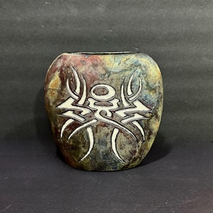 May include: A rounded ceramic vase featuring a mottled, multi-colored glaze in shades of green, brown, and red. The front surface displays a prominent, white, carved tribal-style graphic design with sharp, symmetrical lines and a central oval shape.