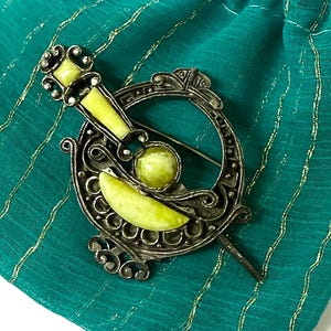 May include: A silver-toned metal brooch featuring an intricate circular frame with scrollwork details. The piece is accented with three smooth, light green stone inlays, including a rectangular top piece, a central sphere, and a curved crescent shape.