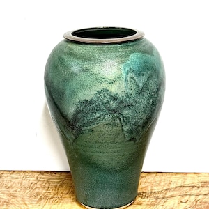 May include: A ceramic vase with a tapered body and a wide opening, featuring a mottled green glaze with darker forest green accents and a smooth, dark brown rim. The surface texture appears matte and slightly granular throughout the exterior.