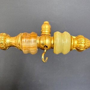 May include: A gold-toned metal rod featuring a decorative end piece with a brown striped stone bead, a central small hook, and a pale green translucent stone bead, finished with a ribbed, pointed finial at the very end of the horizontal structure.