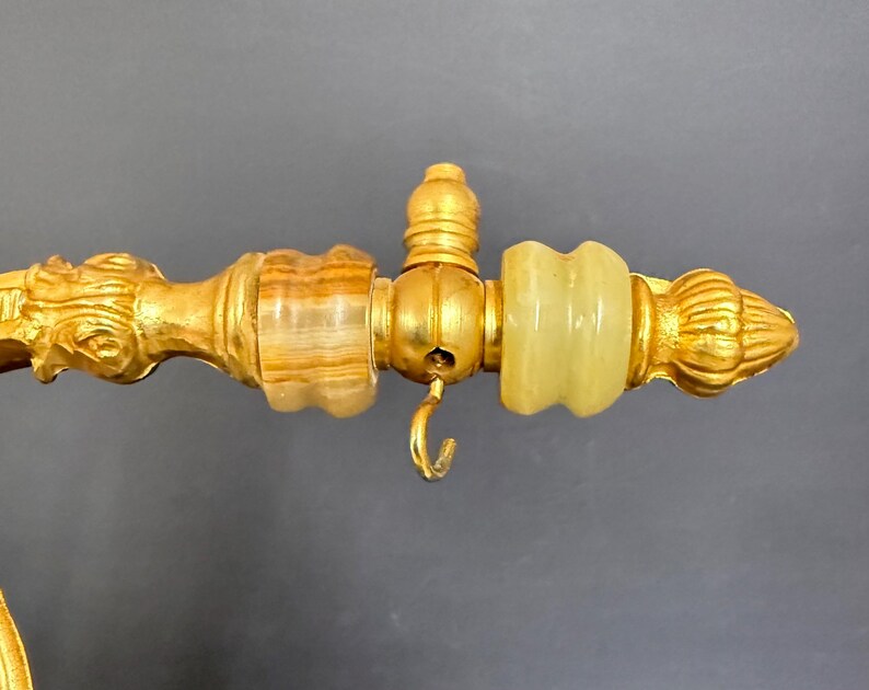May include: A gold-toned metal rod featuring a decorative end piece with a brown striped stone bead, a central small hook, and a pale green translucent stone bead, finished with a ribbed, pointed finial at the very end of the horizontal structure.