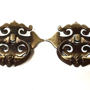 May include: Two antique brass drawer pulls with ornate designs. Each pull features a decorative backplate with scrolled details and a circular handle. The brass has a warm, aged patina, suggesting vintage or retro style.