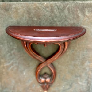 Puede incluir: Una peque&ntilde;a estanter&iacute;a de pared de madera marr&oacute;n oscuro con una parte superior semicircular y un soporte decorativo en forma de coraz&oacute;n. La estanter&iacute;a tiene una ranura en la parte superior y est&aacute; montada sobre una superficie texturizada gris-marr&oacute;n.