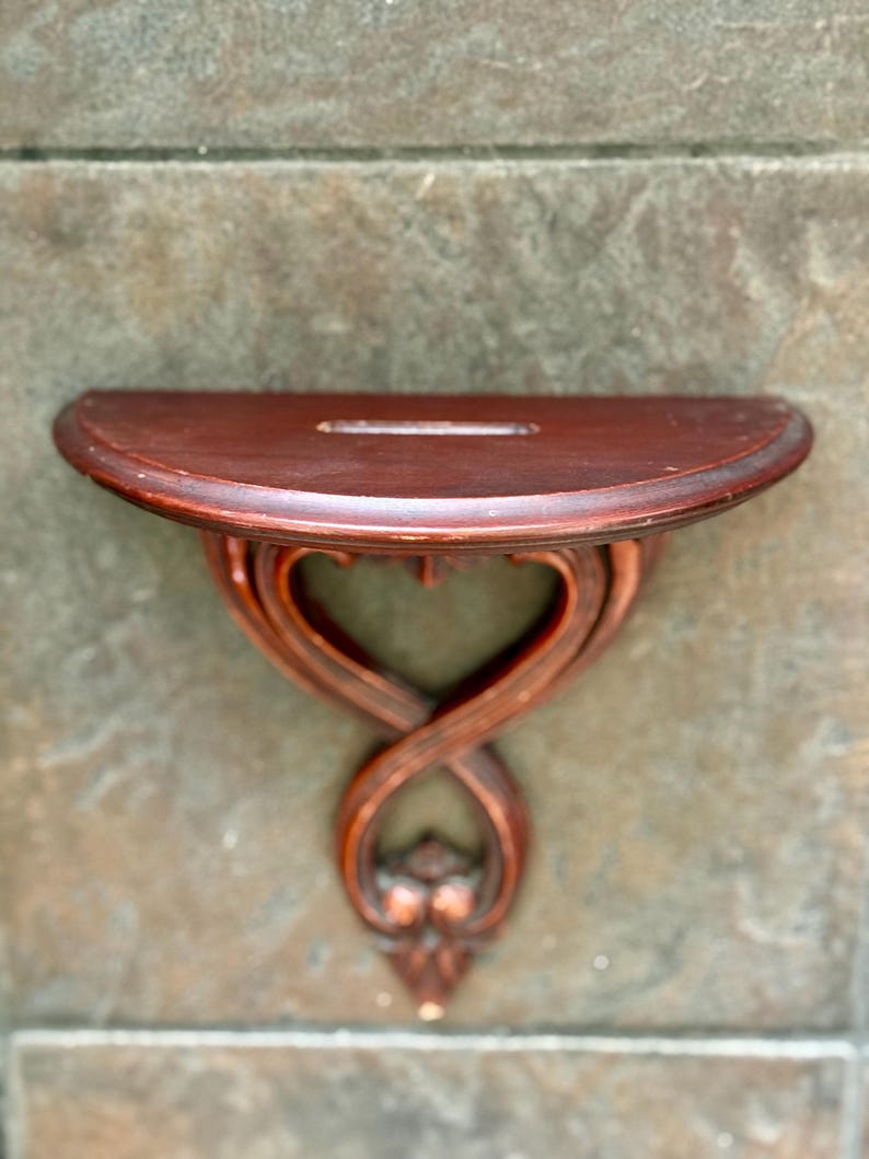 Puede incluir: Una peque&ntilde;a estanter&iacute;a de pared de madera marr&oacute;n oscuro con una parte superior semicircular y un soporte decorativo en forma de coraz&oacute;n. La estanter&iacute;a tiene una ranura en la parte superior y est&aacute; montada sobre una superficie texturizada gris-marr&oacute;n.
