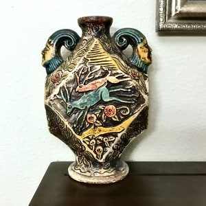 May include: A decorative ceramic vase with a detailed relief design. The vase features a central panel with a deer and floral elements, flanked by handles with human face sculptures. The color palette includes brown, yellow, green, and blue.