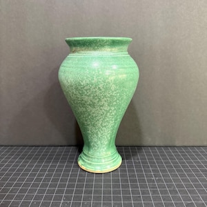 May include: A green ceramic vase with a speckled texture. The vase has a wide, rounded body that tapers towards the base and flares out at the top. The rim is slightly darker green. The vase is approximately 8 inches tall.