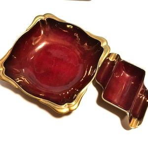 May include: A set of two decorative items: a square-shaped bowl and a smaller ashtray. Both pieces have a deep red interior and gold-colored trim. The bowl has a wavy edge, while the ashtray has a unique, curved design.