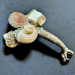May include: Ornate, handcrafted hammer-shaped object. Features a handle with detailed floral patterns and a claw-like end holding a clear sphere. The head incorporates seashells, a square stone, and a crystal. The shells have a spiral design and a pearlescent finish.