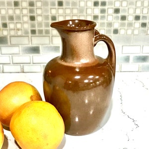 May include: A brown ceramic pitcher with a glossy finish and a curved handle, positioned next to two round, yellow-orange citrus fruits. The pitcher features a tapered neck and a small pouring spout, showcasing a smooth, reflective surface texture.