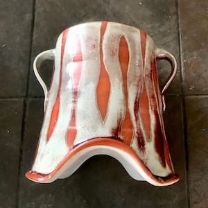 May include: A handmade ceramic vase with a unique shape and two handles. The vase features a white and reddish-orange glaze with vertical, flame-like patterns. The base has a curved, flared design.