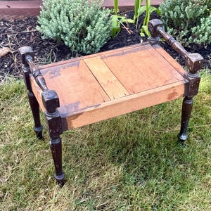 May include: A small wooden footstool with a rectangular top and four dark brown turned legs. The top surface features a light brown wood grain finish with visible signs of wear, while the legs have a dark brown painted finish with scuffs.