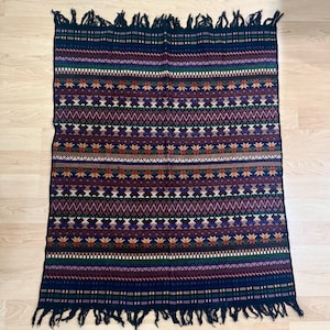 May include: A rectangular woven blanket featuring a dark navy blue base with horizontal bands of intricate geometric patterns in shades of red, green, white, and yellow. The textile includes repeating star and diamond motifs and has fringe.