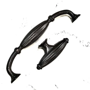 May include: Two dark bronze cabinet pulls. One is a curved handle with a ribbed center and decorative ends. The other is a smaller, matching knob with a similar ribbed design. Both are set against a white background.