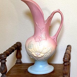 May include: A ceramic pitcher featuring a gradient color scheme that transitions from a soft pink at the top to a pale blue at the base. The body of the pitcher is decorated with a raised, molded floral relief design in cream and green tones.