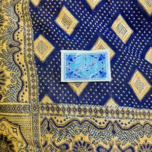 May include: A deep blue fabric featuring a repeating pattern of gold diamonds and small dots, with a wide gold and silver border displaying intricate floral motifs. A rectangular white label with blue text is centered on the fabric.