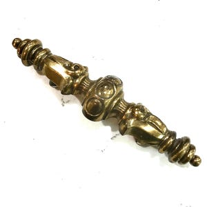 May include: A vintage brass drawer pull with an ornate design. The pull features a central section with three circular details and ends with a spiral design. The brass has a warm, aged patina.