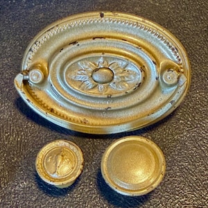 May include: A gold-toned oval drawer pull with a central floral motif and two matching circular knobs, one featuring a small embossed figure and the other a plain smooth surface. All three metal hardware pieces show signs of wear.